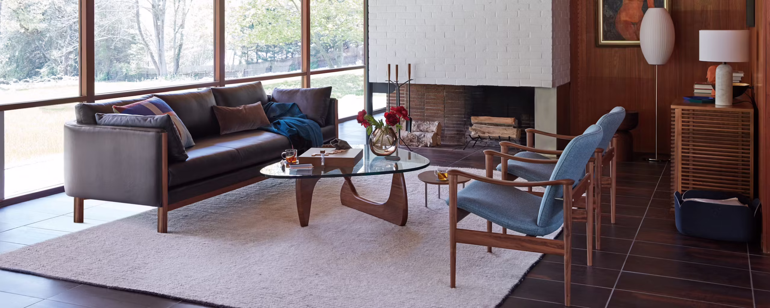 Modern living room featuring a leather sofa, glass coffee table, and two wooden chairs. The room has large windows with a view of nature, a fireplace with a decorative mantel, and a cozy rug.