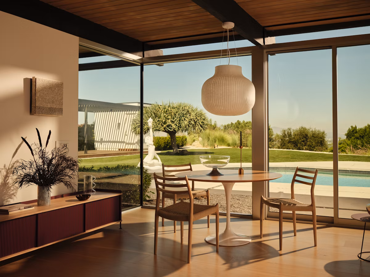 A bright and modern dining space featuring the Knoll Saarinen Round Dining Table with a sculptural base and circular top, surrounded by wooden chairs. Large windows frame a view of a landscaped garden and a pool.